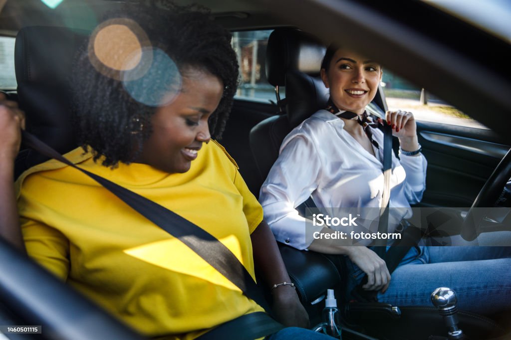 Two girlfriends sitting in a car and fastening their seat belts before going for a drive Candid shot of two diverse young women sitting in a car and fastening their seat belts before going for a drive. Car Pooling Stock Photo
