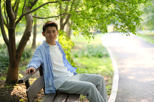 smile handsome Asian young man sit on bench under green tree at park on sunny day. Looking at camera