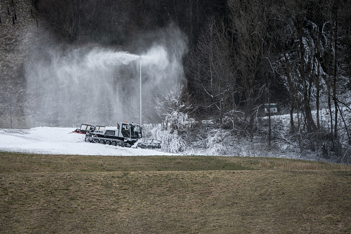 Snow lance cannon producing artificial snow and piste machine prepares the snow in front of brownish green grass as concept for global warming snow shortage winter winter tourism problems