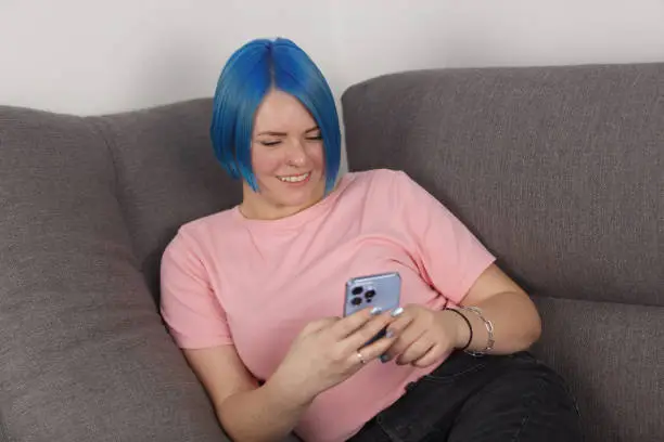 Young adult Ukrainian woman lying on a couch and using modern blue smart phone with a smile