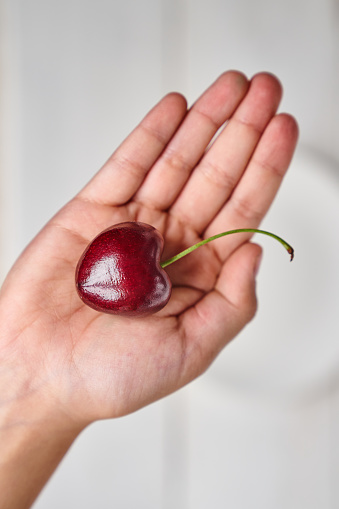 A close-up of a ripe large cherry in the shape of a heart in the hand of a girl, against a white plate. The concept of healthy nutrition and vitamins with space to copy. High quality photo