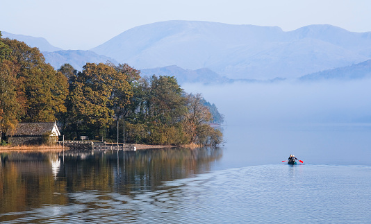 Lake District, UK - October 12, 2010. Boat house on shore of Coniston Water with couple paddling a double canoe. Misty morning in autumn.