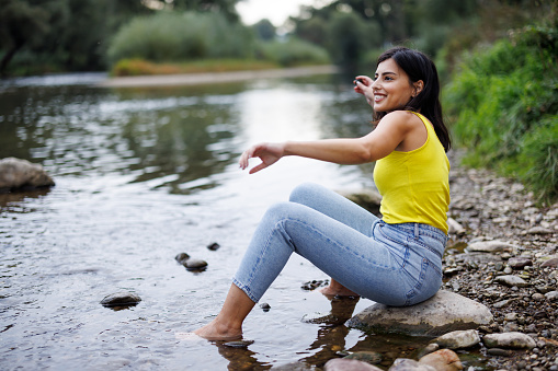 Young woman sitting on rock by a stream with bare feet in water and skimming pebble