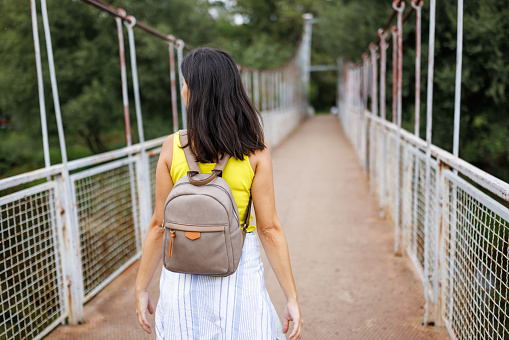 Back view of cheerful young woman walking across footbridge in nature on sunny day