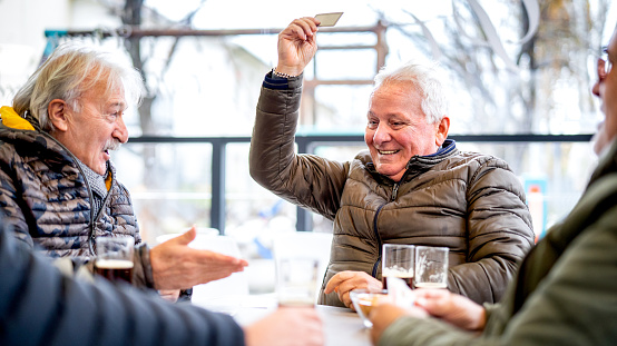 Senior friends playing cards at local bar on winter day - Ageless life style concept with mature people having fun together - Bright contrast filter