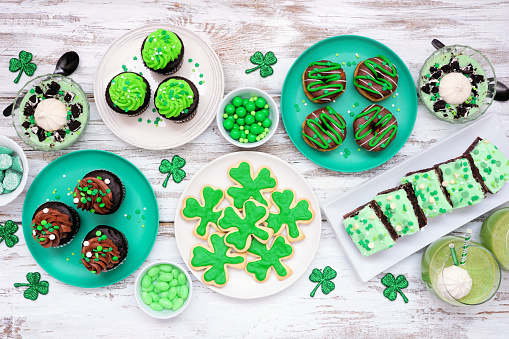 St Patricks Day theme desserts. Table scene over a white wood background. Shamrock cookies, green cupcakes, brownies, donuts and sweets. Overhead view.