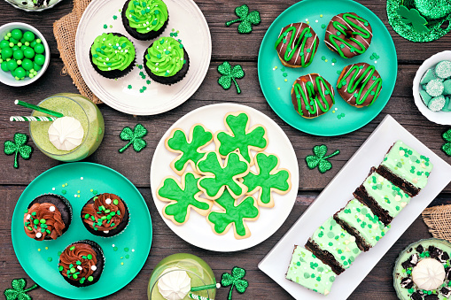 St Patricks Day theme desserts. Table scene over a dark wood background. Shamrock cookies, green cupcakes, brownies, donuts and sweets. Above view.