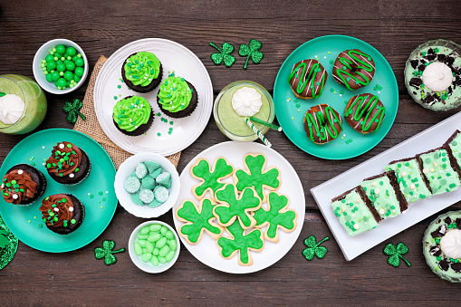 St Patricks Day theme desserts. Table scene over a dark wood background. Shamrock cookies, green cupcakes, brownies, donuts and sweets. Overhead view.