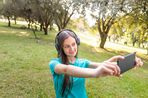 Smiling teenage girl wearing a turquoise t shirt and headphones, taking a selfie with her mobile in a natural space