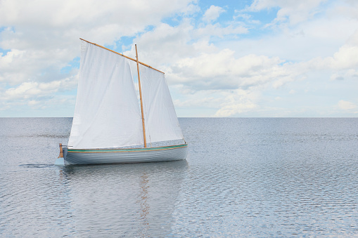 An old boat under a white sail, floating on a calm lake, reflected in the water surface