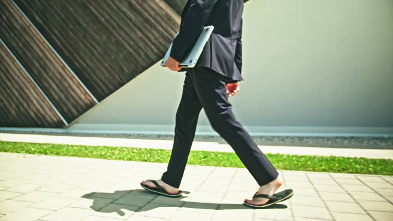 Tracking, slow motion shot of blonde unrecognizable businessman walking, holding a laptop, wearing a black suit and flip flops