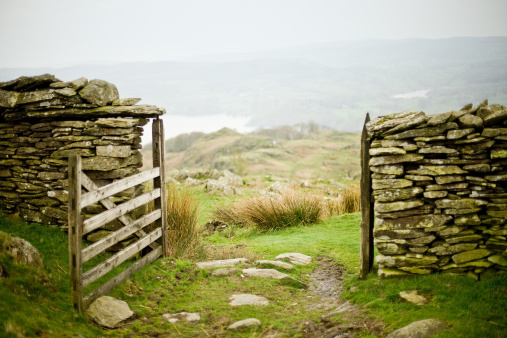 Drystone wall with a farm gate in rural England. The gate is open and leads into a beautiful misty landscape, with green grass and a lake in the background.
