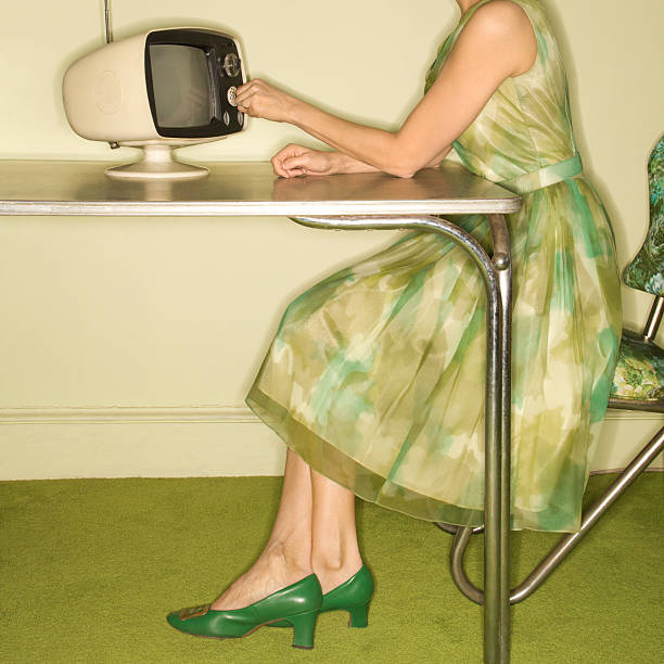 Woman using television. Side view of Caucasian mid-adult woman wearing green vintage dress sitting at 50's retro dinette set turning old televsion knob. 1970s woman stock pictures, royalty-free photos & images