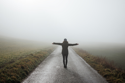 Woman with arms outstretched standing on empty road in fog. Lonely person outdoors