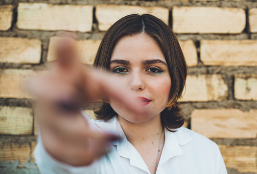 portrait of attractive young woman in white shirt, trying to hold camera with hand, brick wall background.