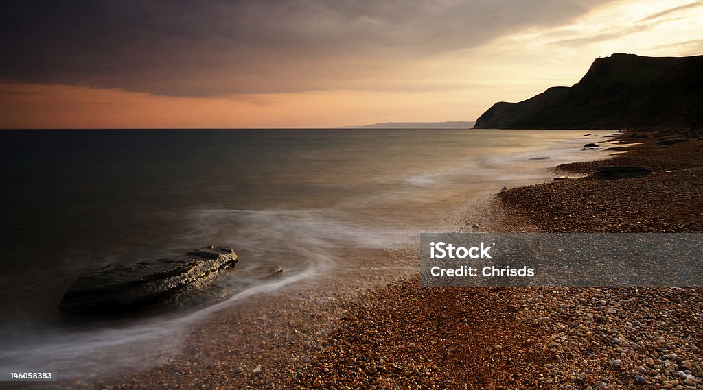 Eype Beach Sunset Dramatic sunset on Eype Beach, Dorset, UK. A long exposure has been used to render the waves as a soft, misty blur or movement. The wet pebble beach reflects the warm evening light and a distant cliff is silhouetted against the light of the setting Summer sun Back Lit Stock Photo