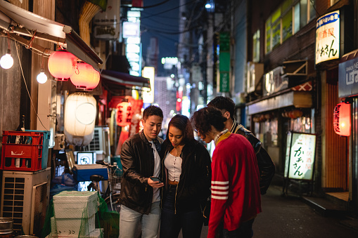 A group of young friends is looking at the cellphone while searching for a place in the phone together to go. They are standing at an illuminated street of Japan at night.