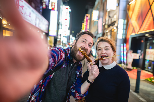 Caucasian couple smiling to the cellphone camera while eating chicken skewers at a street outside in Japan. At the back there are illuminated buildings defocused.