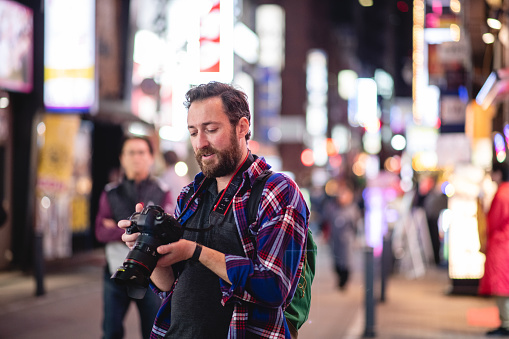 A Caucasian man holds a professional camera and looks at the photos he has taken while he is standing at a crowded street outside in Japan.