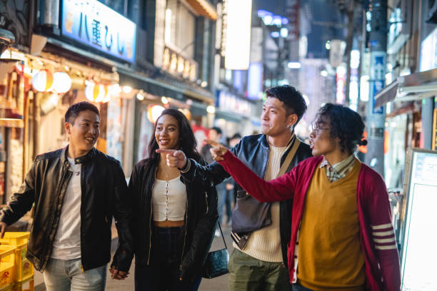 Young Multiracial Friends Pointing a Place While Walking Along An Illuminated Japanese District At Night