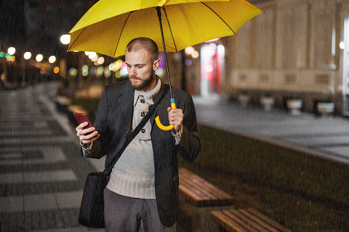 Horizontal medium shot of stylish Caucasian man wearing eyeglasses standing outdoors under transparent umbrella holding coffee cup, looking away and smiling