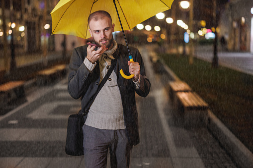 A young man with a yellow umbrella is walking down the city district on a rainy night and talking using a smartphone.