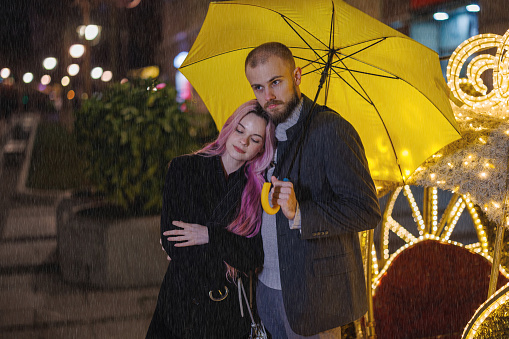 A young man and his girlfriend with a yellow umbrella are walking down the city streets during a rainy autumn night.