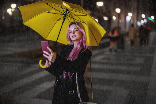 A young woman with a yellow umbrella is walking down the city district on a rainy night and using a smartphone to take selfie photos.