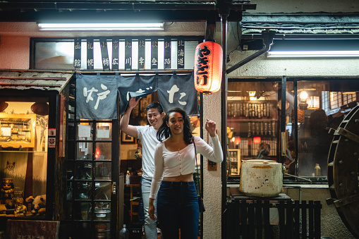 A young multiracial couple is leaving a Japanese bar while raising their hands. They look happy and relaxed while going outdoors.