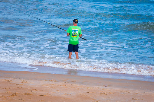 Ormand Beach, Florida, USA-January 3, 2023-  A mature man, wearing a hat, sunglasses and green fishing t-shirt fishes in the ocean while standing calf deep in the surf on an early January afternoon.