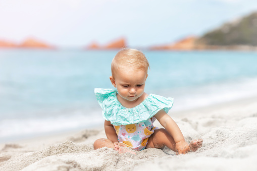 Beautiful baby girl playing with sand while sitting in a swimsuit on the beach in summer. Summer weekend, family travel, vacation concept