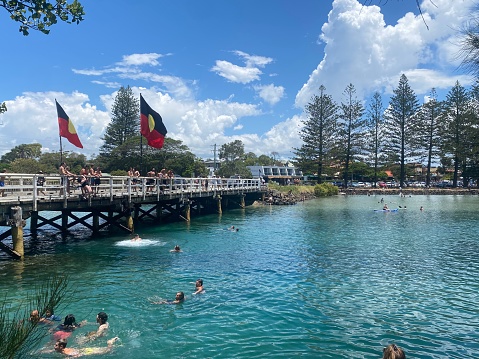 Horizontal seascape of people swimming and enjoying summer in turquoise lake waters under bridge with Aborigional flags flying on Australia Day with coastal features of pine trees and mangrove shoreline under cloudscape at Brunswick Heads NSW Australia