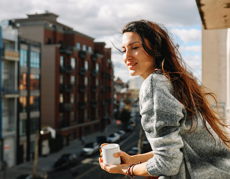 Young woman having a coffee, relaxing at the balcony, enjoying the view in Brooklyn, New York on a bright and sunny day.