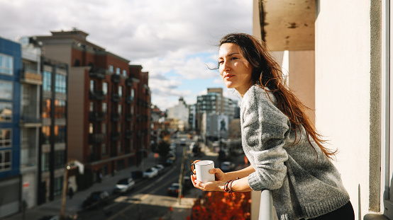 Young woman having a coffee, relaxing at the balcony, enjoying the view in Brooklyn, New York on a bright and sunny day.
