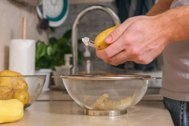 un homme épluche des pommes de terre avec un éplucheur de légumes dans une cuisine lumineuse. préparation des pommes de terre pour la cuisson. nettoyage de la peau des pesticides. un homme prépare le dîner ou le déjeuner à la maison pour la famille - root vegetable raw potato human skin root photos et images de collection