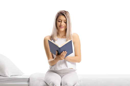 Woman in pajamas sitting on a bed and reading a book isolated on white background