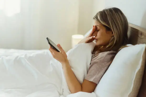 Young woman with a headache using smart phone while relaxing in a bed during morning time. Copy space.