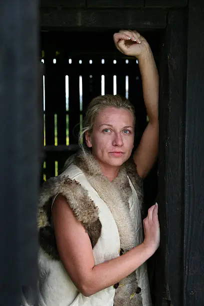 Blond caucasian cowgirl woman leaning in old wood barn doorway, with blue eyes, wearing leather/fur vest.