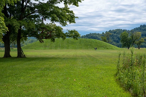 Native American Hopewell Culture prehistoric Seip Earthworks burial mounds in Ohio. Ancient large long mound. Two people read sign in the distance. Grass is neatly trimmed with trees and dramatic sky. Copy space
