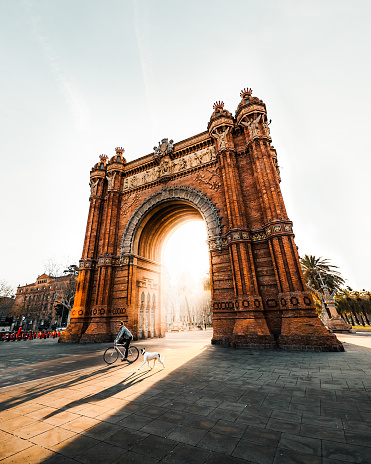 Barcelona, Spain – September 09, 2021: A person riding a bike and a dog passing by the Arc de Triomf in Barcelona, Spain