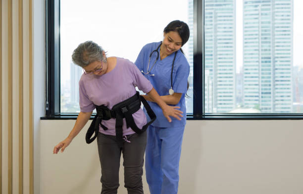 Woman physical therapist in blue uniform help an elderly to exercise and practice walking with Safety Transfer Belt. Atmosphere in Community Health Center.