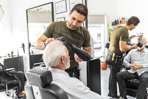 Senior man getting groomed at hairdresser with hair dryer while sitting in chair