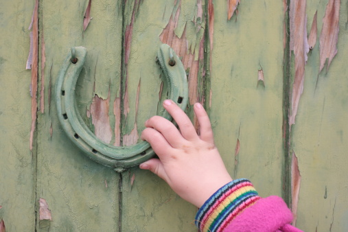 Child's hand using a horse shoe door knocker on a green door.