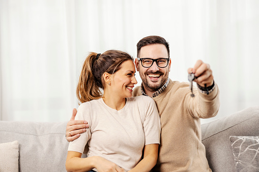 A happy man is hugging his wife at home showing keys at the camera while sitting in a living room.