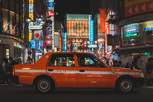 To, Japan – October 29, 2018: An image of an orange taxi with building and neon lights on a background and people walking around in Tokyo, Japan