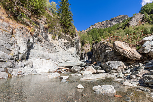 Pond with crystal clear water and small pebbles formed by Lillaz waterfall (Cascate di Lillaz) and hiking trail with wooden railings along crack covered with green forest, Aosta valley, Italy