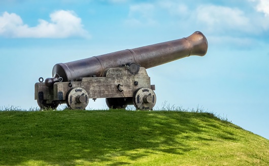 A Cannon on the grassy hill, under the shadow of a tree, with a clouds in the sky on a sunny day