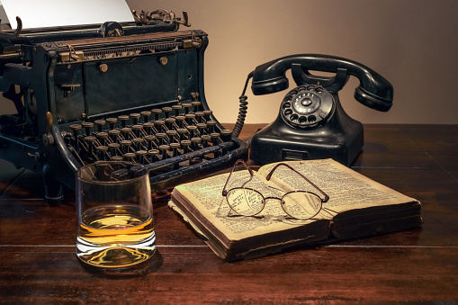 A vintage typewriter and a telephone on the table next to a glass of whiskey and an opened book.