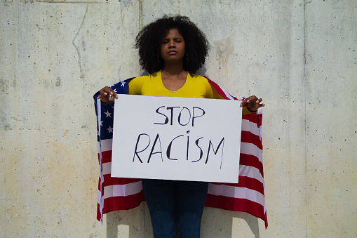 An african-American woman holds a stop racism banner in her hands and united states flag over her shoulders. In background grey wall