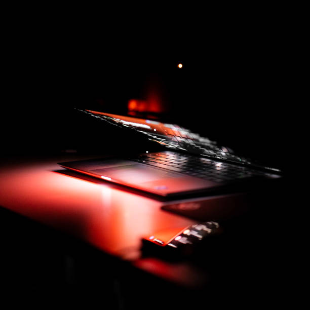 The image shows An Afro Caribbean man using a silver laptop in the dark with the whole scene being lit by the white computer screen. - You only see the mans hands on the keyboard with black keys shot from over his shoulder also you can see the top of the mans jeans and a silver belt buckle.